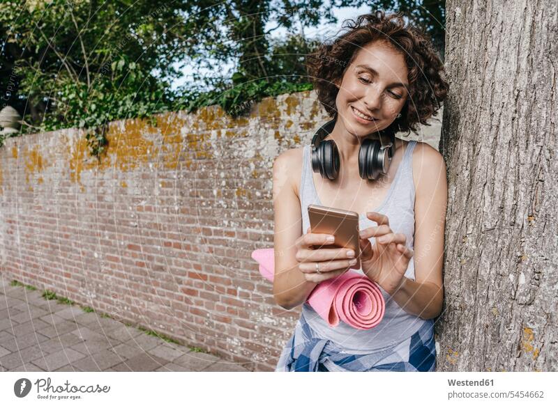 Lächelnde Frau mit Matte, Kopfhörer und Handy auf dem Bürgersteig Mobiltelefon Handies Handys Mobiltelefone weiblich Frauen Telefon telefonieren Kommunikation