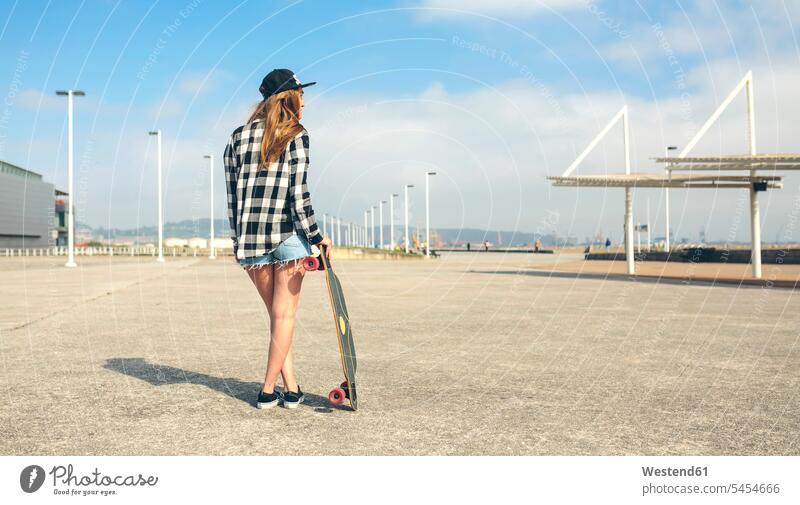 Rückenansicht einer jungen Frau mit Longboard, die vor der Strandpromenade steht, Teilansicht weiblich Frauen Erwachsener erwachsen Mensch Menschen Leute People