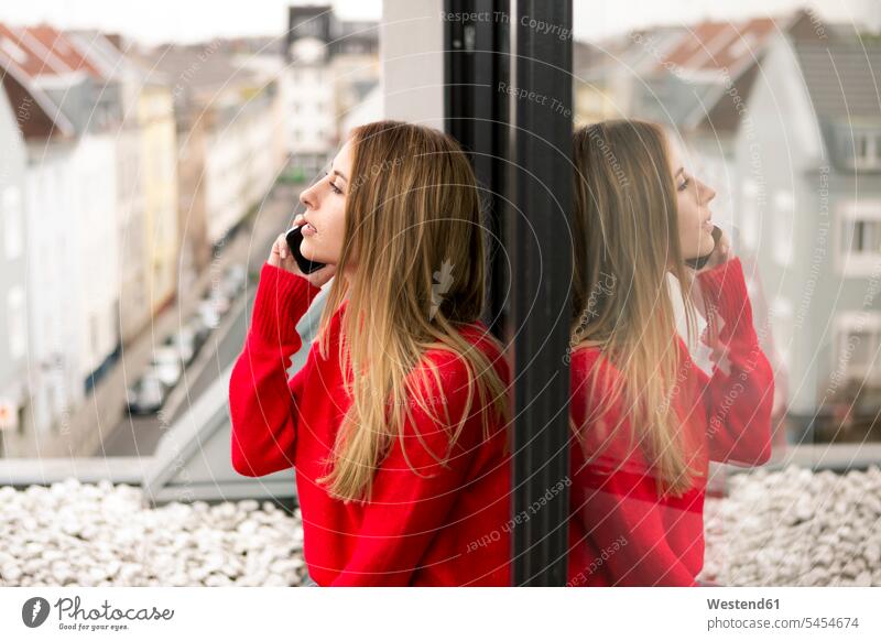 Junge Frau am Fenster in Stadtwohnung am Handy Mobiltelefon Handies Handys Mobiltelefone weiblich Frauen telefonieren anrufen Anruf telephonieren staedtisch