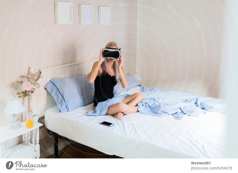 Junge Frau sitzt im Bett und trägt eine VR-Brille Virtuelle Realität Virtuelle Realitaet weiblich Frauen Brillen sitzen sitzend Betten Erwachsener erwachsen