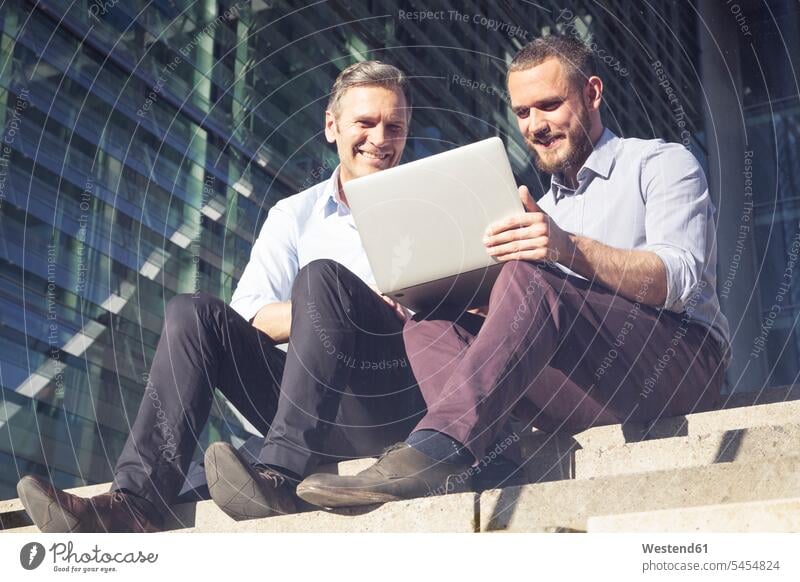 Zwei lächelnde Geschäftsleute sitzen mit Laptop auf der Treppe Treppenaufgang Notebook Laptops Notebooks sitzend sitzt Kollegen Arbeitskollegen Geschäftsmann