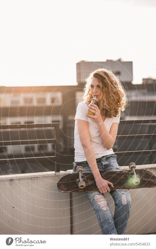 Junge Frau mit Skateboard trinkt Getränk auf der Dachterrasse bei Sonnenuntergang Sonnenuntergänge Rollbretter Skateboards weiblich Frauen Getraenk Getränke