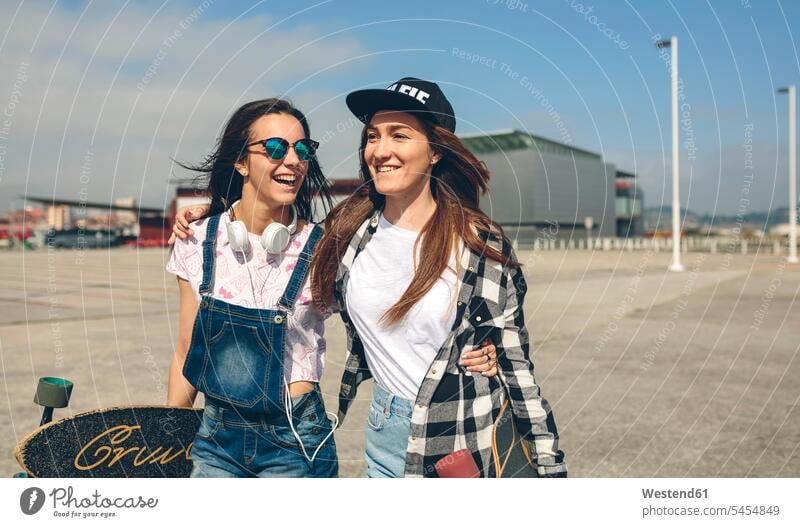 Zwei glückliche junge Frauen mit Longboards gehen Arm in Arm an der Strandpromenade Freundinnen Freunde Freundschaft Kameradschaft weiblich lachen Skateboard