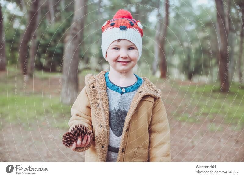 Bildnis eines Jungen mit Wollhut, der im Wald Kiefernzapfen hält lächeln Wollmütze Wollmützen Strickmütze Strickmützen Buben Knabe Knaben männlich Zapfen Mütze