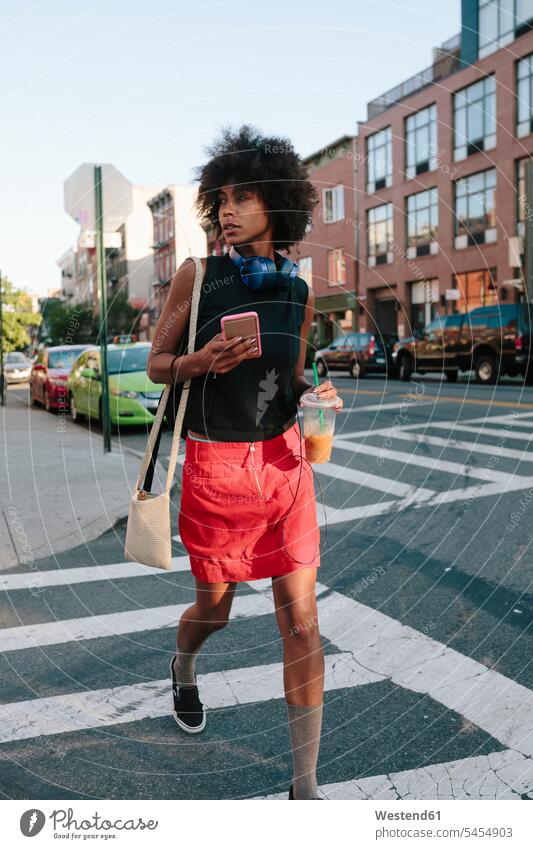 Junge Frau mit Kopfhörern und Smartphone auf der Querstraße in Brooklyn, die ein Getränk zum Mitnehmen anbietet gehen gehend geht Handy Mobiltelefon Handies