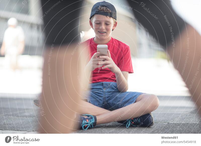 Junge benutzt Smartphone, sitzt auf Skateboard Handy Mobiltelefon Handies Handys Mobiltelefone lesen Lektüre simsen sitzen sitzend Rollbretter Skateboards SMS