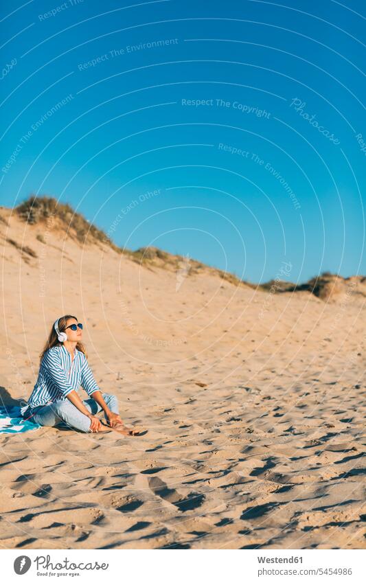 Portugal, Aveiro, Frau sitzt in der Nähe einer Stranddüne und hört Musik mit Kopfhörern Strandduene Strandduenen Stranddünen weiblich Frauen Sand sandig