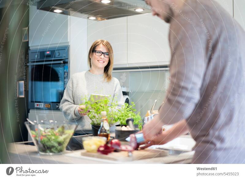 Lächelnde Frau mit Tablette sieht ihren Freund an, der in der Küche Salat zubereitet Paar Pärchen Paare Partnerschaft lächeln Tablet Computer Tablet-PC