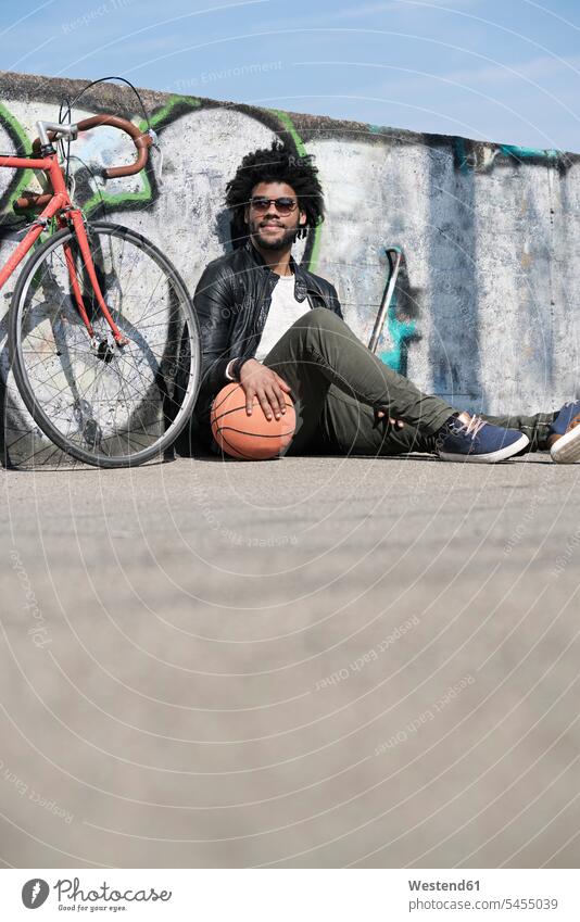 Lächelnder Mann sitzt vor der Grafitti-Wand neben seinem Fahrrad und hält Basketball Männer männlich lächeln Bikes Fahrräder Räder Rad sitzen sitzend