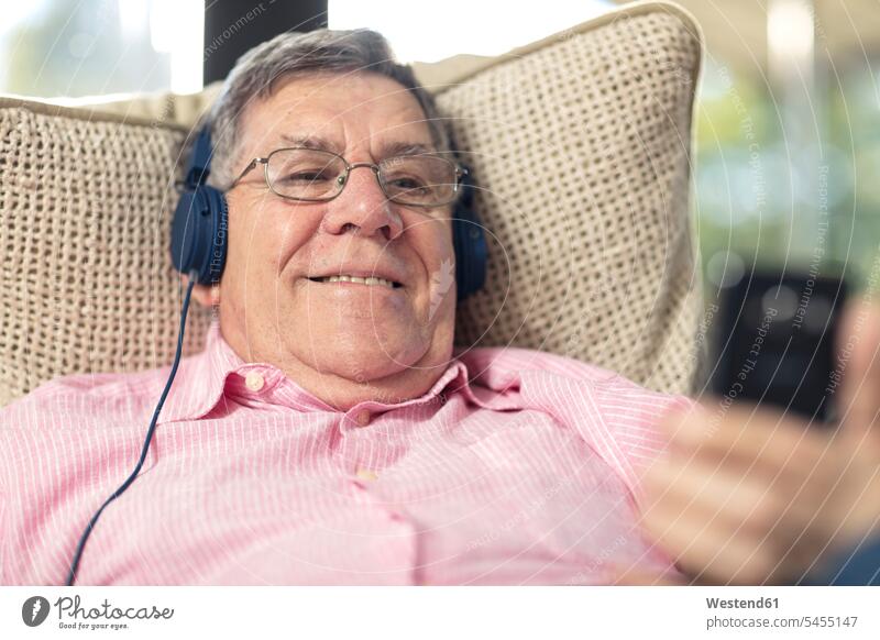Lächelnder älterer Mann mit Kopfhörern, der zu Hause Musik hört Zuhause daheim lächeln Männer männlich hören hoeren Sofa Couches Liege Sofas Kopfhoerer Senior