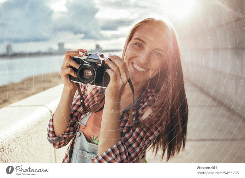 Lächelnde junge Frau mit einer Kamera am Flussufer weiblich Frauen Fotoapparat Fotokamera Portrait Porträts Portraits lächeln Fluesse Fluß Flüsse Erwachsener