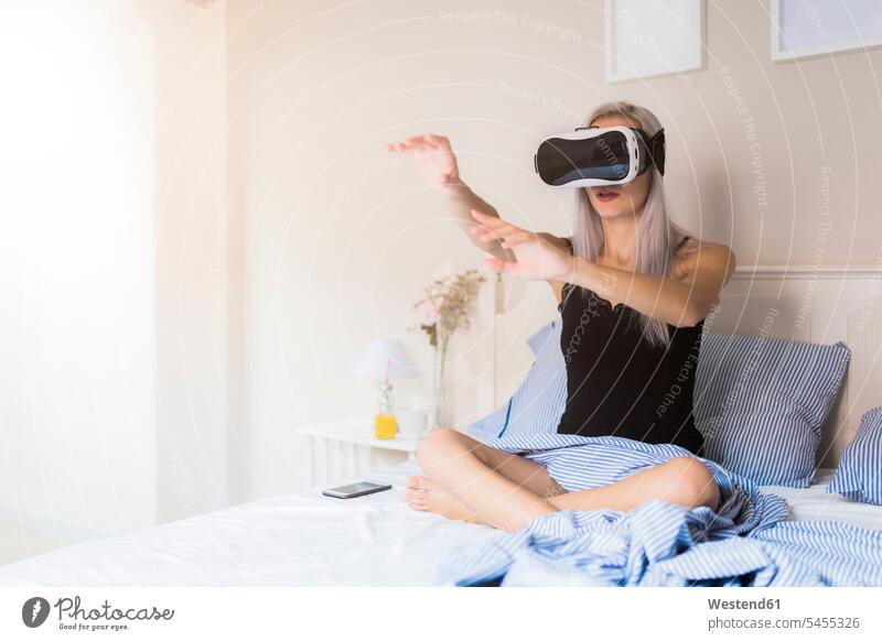 Junge Frau sitzt im Bett und trägt eine VR-Brille Virtuelle Realität Virtuelle Realitaet Brillen Betten weiblich Frauen sitzen sitzend Erwachsener erwachsen