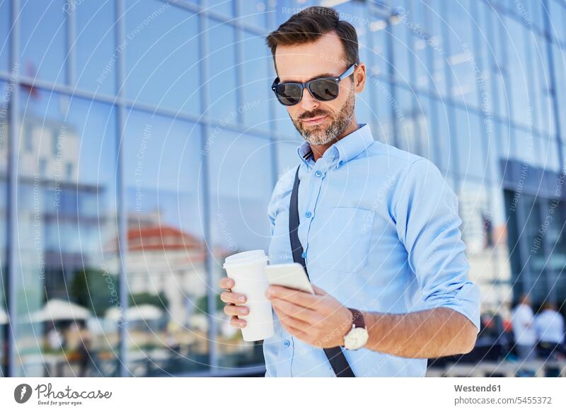 Geschäftsmann benutzt Telefon und hält Kaffee im Freien stehen stehend steht Handy Mobiltelefon Handies Handys Mobiltelefone Businessmann Businessmänner