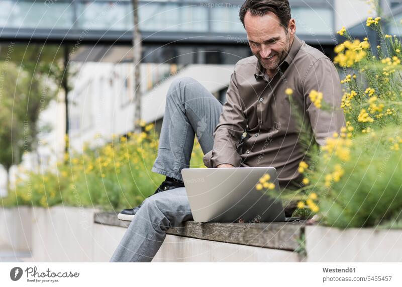 Lächelnder Mann sitzt mit Laptop an der Wand Geschäftsmann Businessmann Businessmänner Geschäftsmänner Freiberufler freiberuflich freie Berufe Notebook Laptops