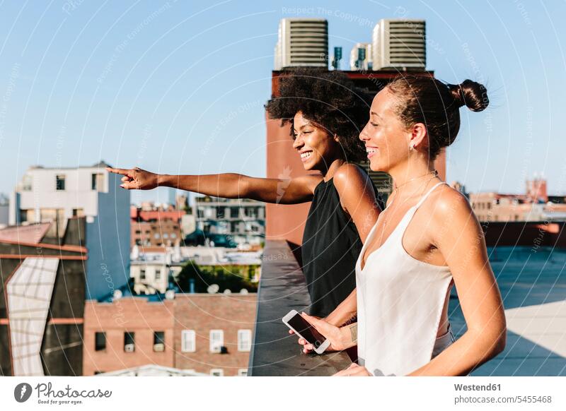 Freundinnen, die auf dem Dach stehen und in die Ferne zeigen lächeln Freude freuen Dachterrasse Dachterrassen Smartphone iPhone Smartphones Unbeschwert Sorglos