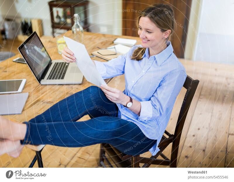 Lächelnde Frau sitzt zu Hause am Schreibtisch mit den Füssen nach oben und liest Dokument Zuhause daheim sitzen sitzend lesen Lektüre weiblich Frauen Unterlagen