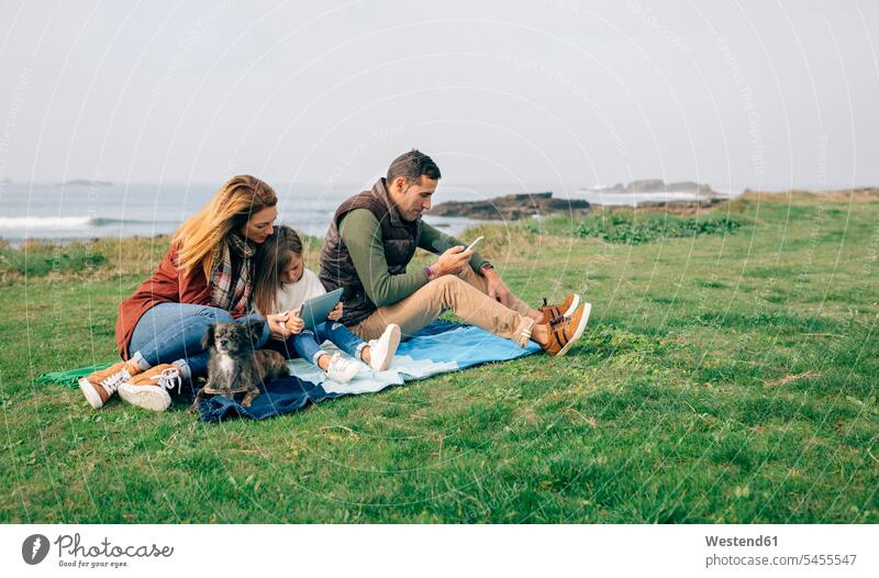 Familie mit Hund sitzt mit drahtlosen Geräten auf einer Decke an der Küste Hunde Kueste Kuesten Küsten Decken Familien Haustier Haustiere Tier Tierwelt Tiere