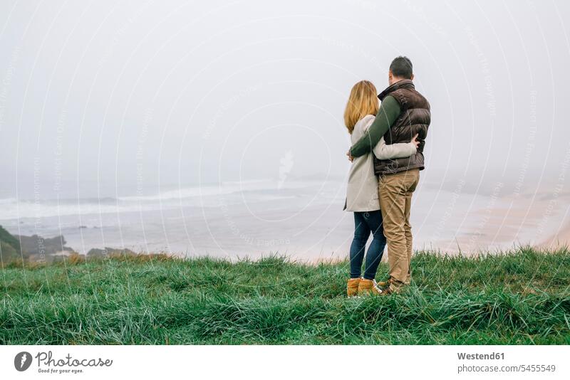 Paar, das sich an einem nebligen Wintertag an der Küste umarmt und die Aussicht betrachtet umarmen Umarmung Umarmungen Arm umlegen Pärchen Paare Partnerschaft