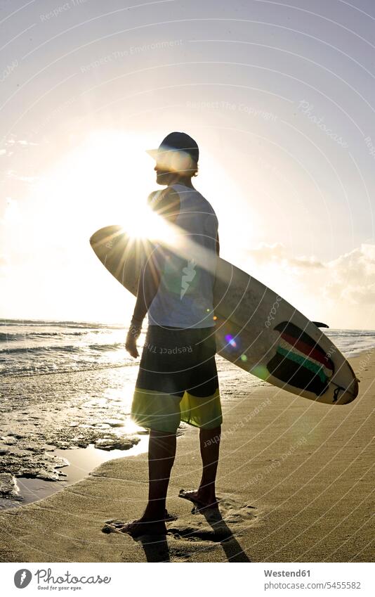 Mann mit Surfbrett steht am Strand und sieht den Sonnenuntergang Beach Straende Strände Beaches Männer männlich Erwachsener erwachsen Mensch Menschen Leute