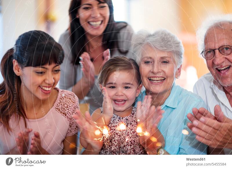Kleines Mädchen schaut sich Wunderkerzen auf einer Geburtstagstorte an, sitzt auf dem Schoß der Großmutter, die Familie ist dabei Geburtstagskuchen