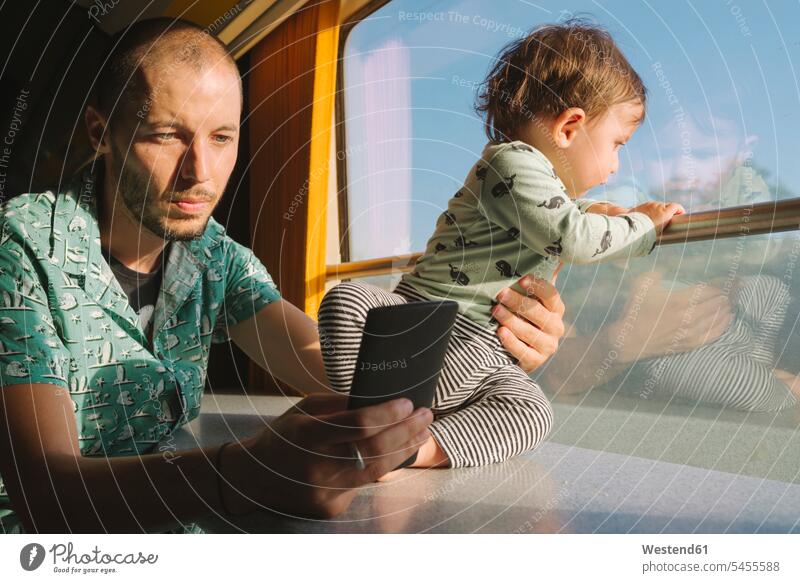 Vater hält sein kleines Mädchen und benutzt ein Smartphone, während er mit dem Zug reist Papas Väter Vati Vatis Papis Bahnen Züge reisen verreisen Handy