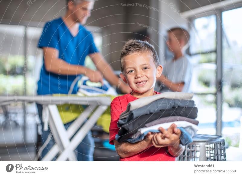 Lächelnder Junge hilft bei der Hausarbeit und hält gefaltete Kleidung tragen transportieren Hausarbeiten lächeln helfen mithelfen Hilfsbereitschaft beistehen