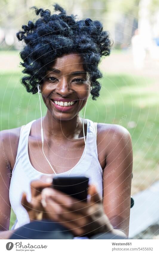 Porträt einer lächelnden Frau mit Handy und Kopfhörer in einem Park weiblich Frauen Smartphone iPhone Smartphones Portrait Porträts Portraits Erwachsener
