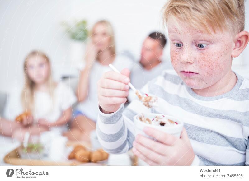 Junge starrt auf Müslischale mit Familie im Hintergrund essen essend Buben Knabe Jungen Knaben männlich Kind Kinder Kids Mensch Menschen Leute People Personen