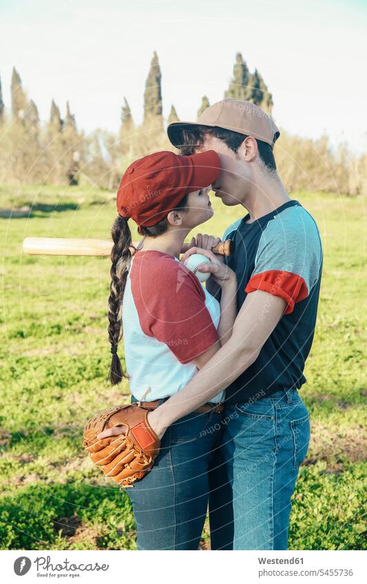 Junges Paar mit Baseballausrüstung küsst sich im Park Baseballspiel Baseballspieler Baseballer Pärchen Paare Partnerschaft küssen Küsse Kuss Sport Mensch