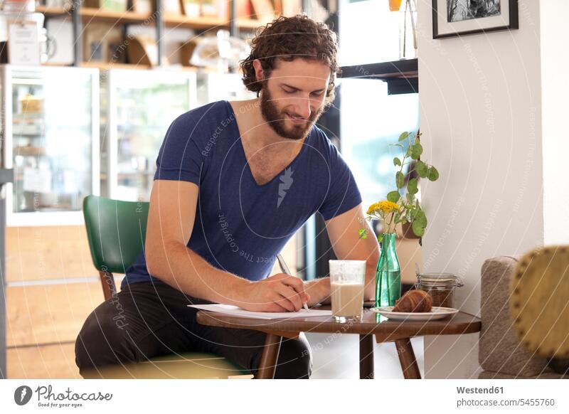 Lächelnder junger Mann sitzt in einem Café und schreibt einen Brief schreiben aufschreiben notieren schreibend Schrift Männer männlich Cafe Kaffeehaus Bistro