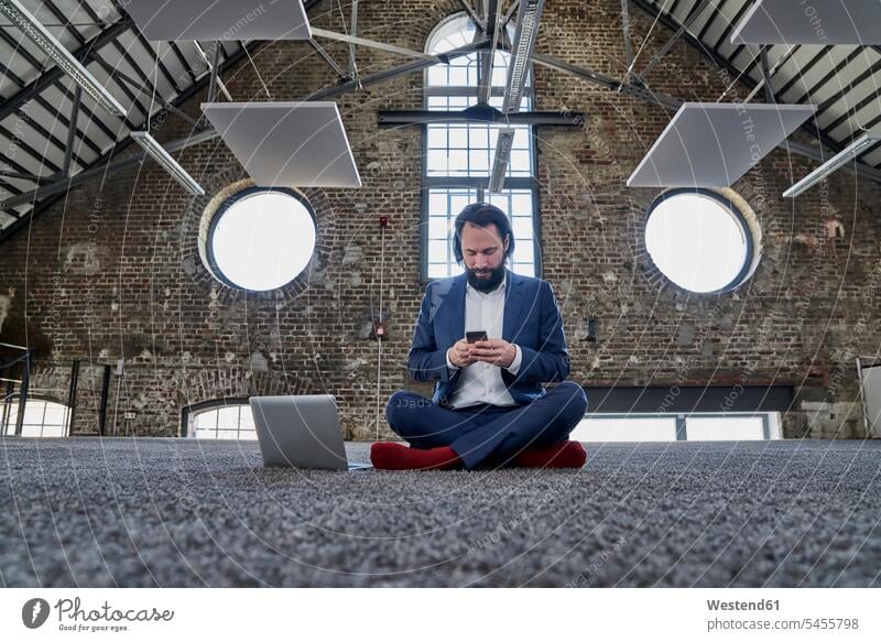 Geschäftsmann sitzt auf einem Teppich in einem Loft und benutzt sein Handy Businessmann Businessmänner Geschäftsmänner Mobiltelefon Handies Handys Mobiltelefone