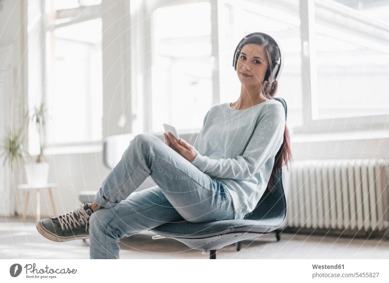 Junge Frau, die von ihrem Mobiltelefon aus einen Videoanruf macht und Kopfhörer trägt weiblich Frauen Kopfhoerer Stuhl Stuehle Stühle Handy Handies Handys