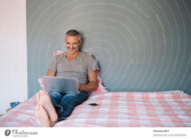 Lächelnder reifer Mann sitzt zu Hause auf dem Bett und benutzt einen Laptop Notebook Laptops Notebooks Zuhause daheim Männer männlich benutzen lächeln sitzen