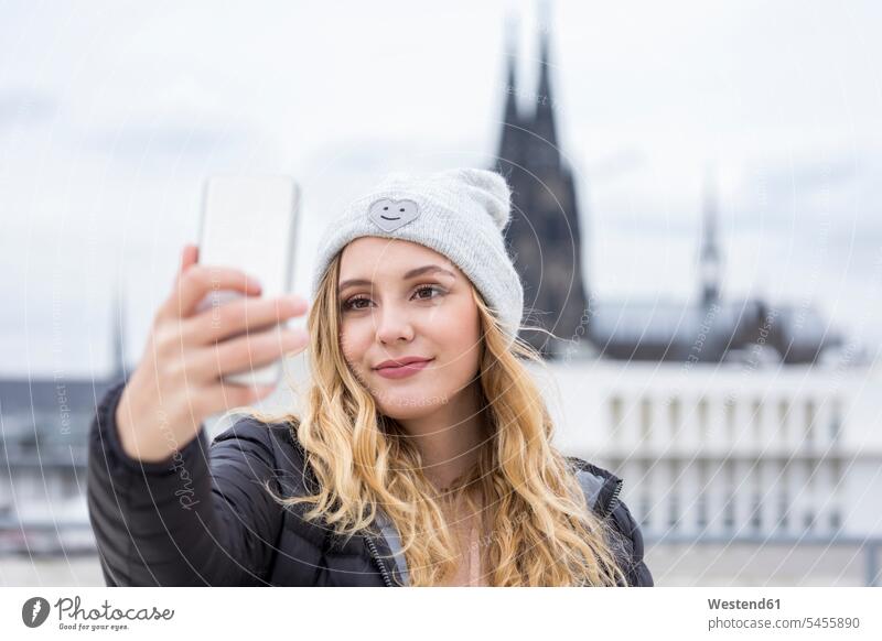 Deutschland, Köln, Porträt einer lächelnden jungen Frau, die ein Selfie mit ihrem Smartphone macht Portrait Porträts Portraits Selfies weiblich Frauen iPhone