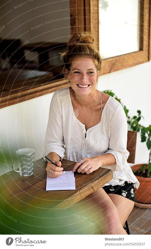 Porträt einer lachenden jungen Frau mit Tasse Kaffee, die auf der Veranda eines Cafés sitzt und schreibt Portrait Porträts Portraits weiblich Frauen Erwachsener