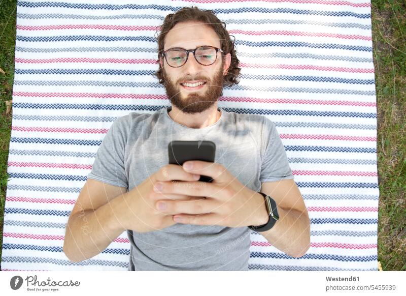 Porträt eines lächelnden Mannes, der in einem Park auf einer Decke liegt und sein Handy benutzt, Draufsicht Portrait Porträts Portraits Männer männlich