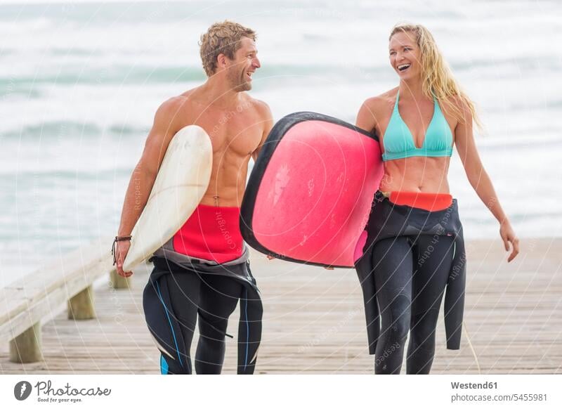 Paare, die mit Surfbrettern vom Strand aus laufen Beach Straende Strände Beaches surfboard surfboards Surfen Surfing Wellenreiten Pärchen Partnerschaft lachen