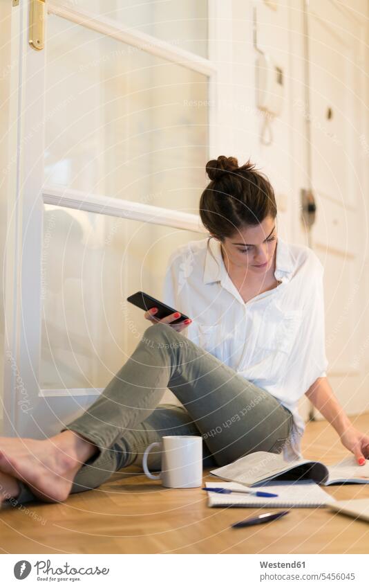 Junge Frau sitzt auf dem Boden mit Kaffeetasse und Smartphone und betrachtet Broschüre weiblich Frauen Erwachsener erwachsen Mensch Menschen Leute People