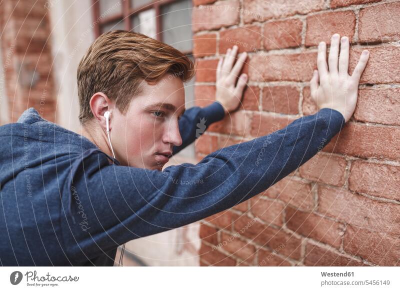 Junger Mann mit Kopfhörern an Backsteingebäude gelehnt Männer männlich Ohrhörer ernst Ernst Ernsthaftigkeit ernsthaft hören hoeren Erwachsener erwachsen Mensch