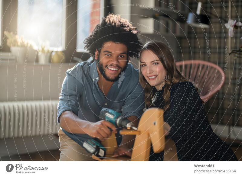 Lächelndes Paar schaut auf eine Kamera, die ein Schaukelpferd mit Akkubohrer montiert Zuhause zu Hause daheim Pärchen Paare Partnerschaft lächeln Schaukelpferde