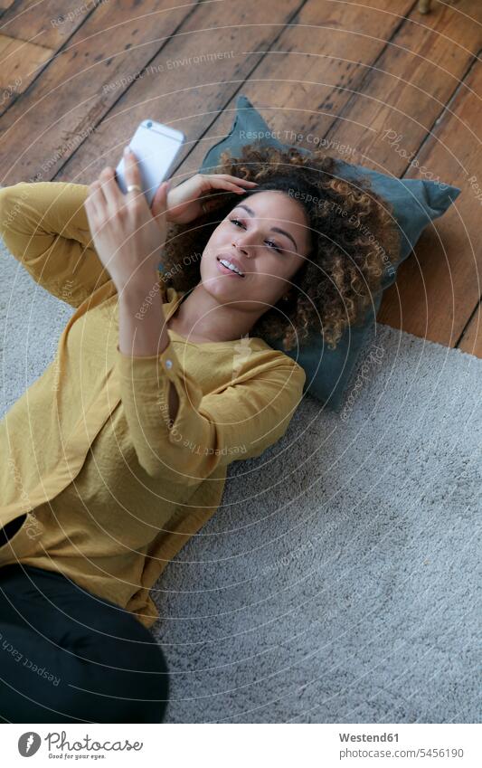 Lächelnde junge Frau liegt zu Hause mit Handy auf dem Boden Zuhause daheim lächeln liegen liegend Böden Boeden Mobiltelefon Handies Handys Mobiltelefone
