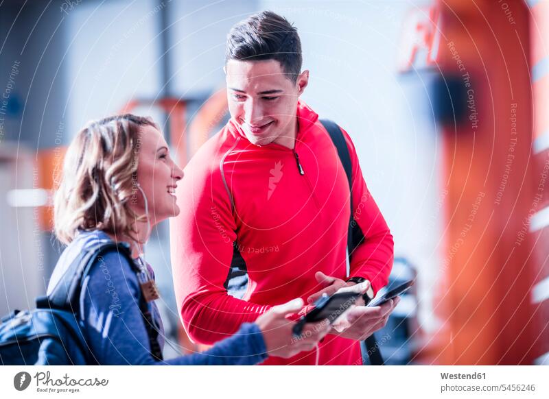 Junger Mann und Frau mit Mobiltelefonen und Ohrstöpseln im Fitnessstudio lächeln Pause trainieren Fitnessclubs Fitnessstudios Turnhalle fit Gesundheit gesund