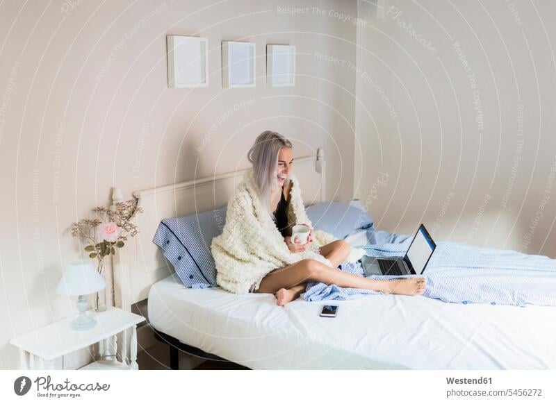Glückliche junge Frau im Bett mit Tasse Kaffee und Laptop glücklich glücklich sein glücklichsein Kaffeetasse Kaffeetassen Notebook Laptops Notebooks Betten
