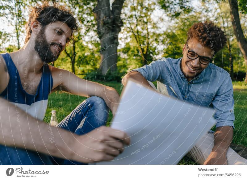 Zwei Freunde sitzen in einem Park und diskutieren über Papiere sitzend sitzt Parkanlagen Parks Unterlagen besprechen Besprechung Mann Männer männlich