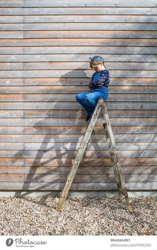 Mädchen sitzt auf einer Leiter vor einer Holzfassade und schmollt mit dem Mund Leitern sitzen sitzend schmollen schmollend weiblich Kind Kinder Kids Mensch