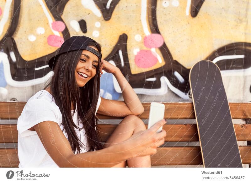 Lächelnde junge Frau mit Skateboard schaut auf Handy Mobiltelefon Handies Handys Mobiltelefone weiblich Frauen Bank Sitzbänke Bänke Sitzbank lachen Rollbretter