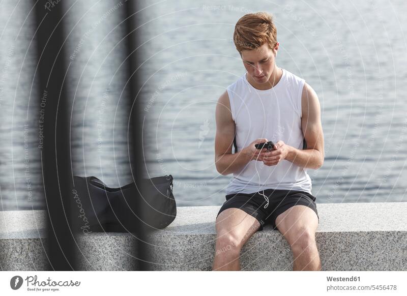 Junger Sportler sitzt mit Handy und Kopfhörer auf einer Mauer am Flussufer trainieren Joggen Jogging Mauern Tasche Taschen Mobiltelefon Handies Handys