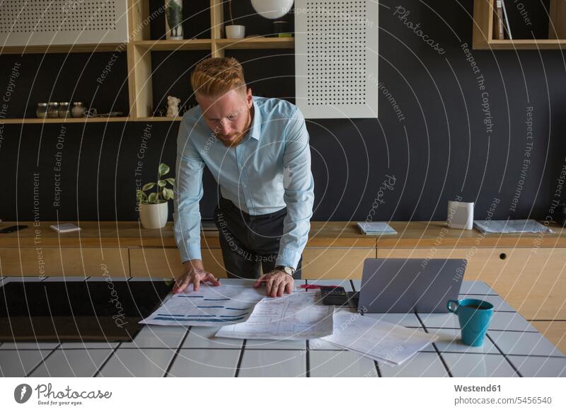 Mann mit Laptop studiert Pläne auf dem Tisch zu Hause Zuhause Plan studieren Computer Erwachsener Mensch Kaffeebecher betrachten Bart Mobilität Kalkulation