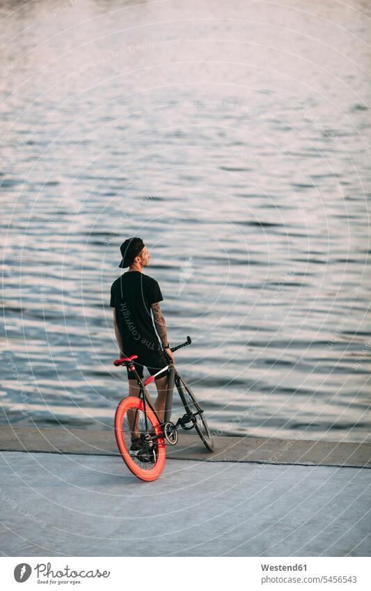 Junger Mann mit Fixie-Rad am Wasser Fluss Fluesse Fluß Flüsse stehen stehend steht Fahrrad Bikes Fahrräder Räder Männer männlich Gewässer Raeder Erwachsener