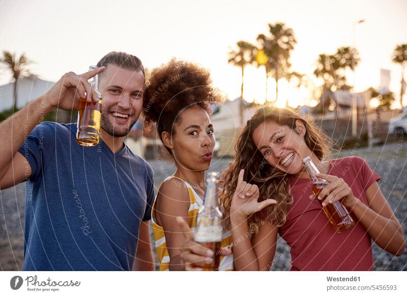 Drei Freunde mit Bierflaschen, die sich bei Sonnenuntergang am Strand amüsieren Beach Straende Strände Beaches Alkohol Alkoholische Getraenke